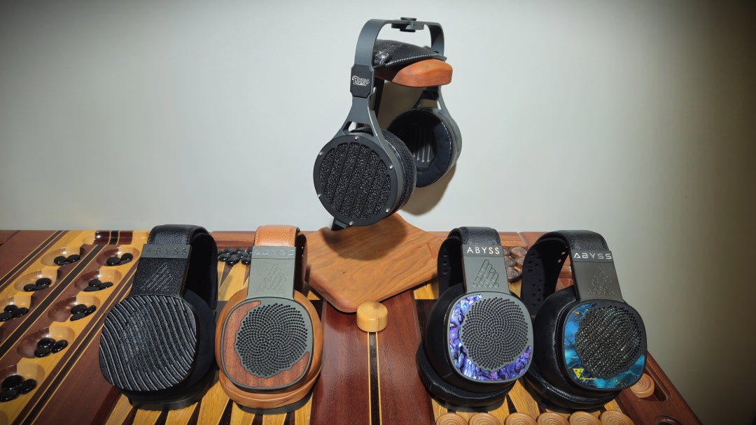 Four Abyss over-ear headphones displayed on a wooden surface against a plain wall, with one pair hanging on a wooden stand above the others.